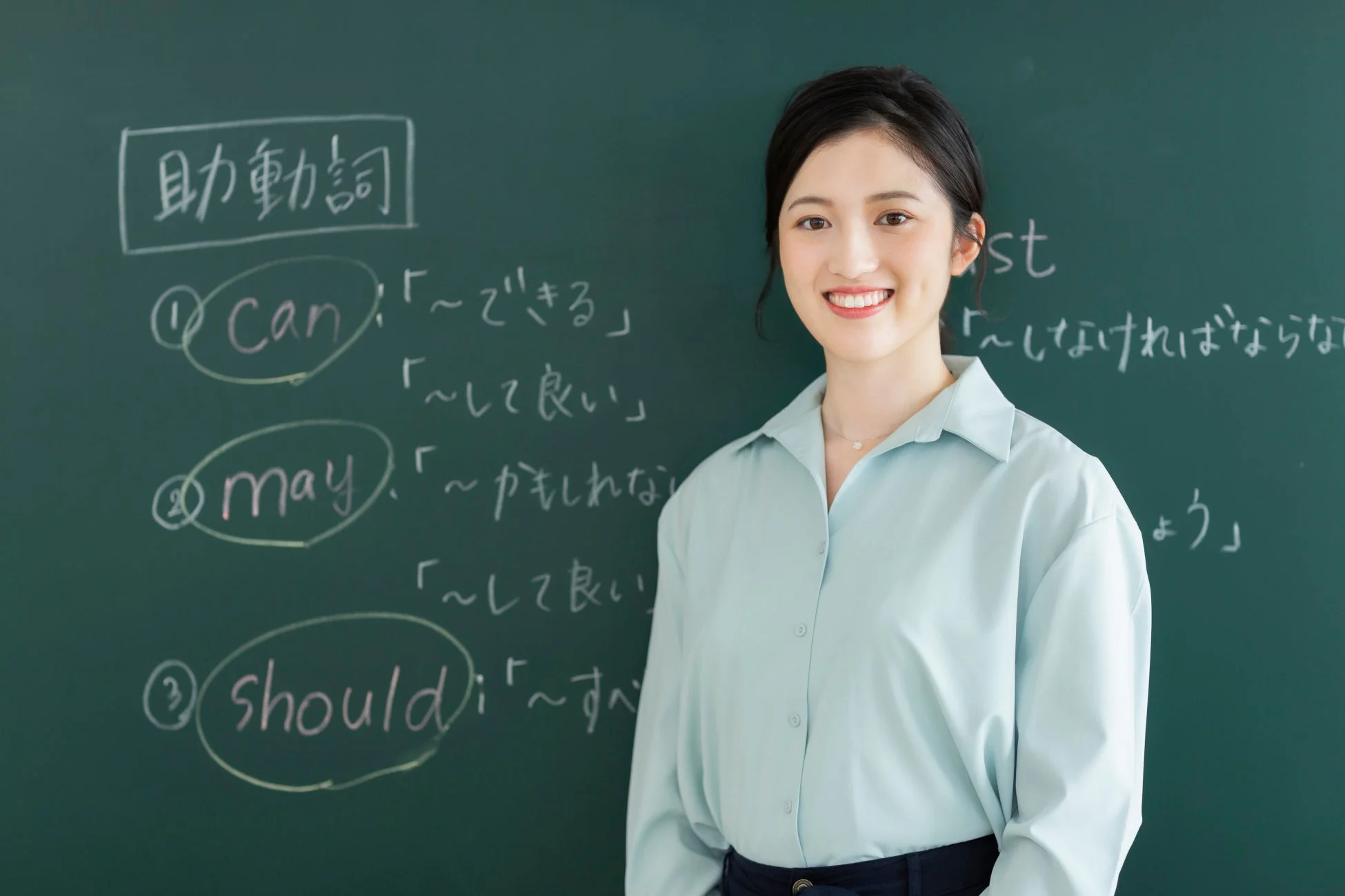 笑顔の若い女性が黒板の前に立っており、英語の助動詞とその意味が書かれた授業風景