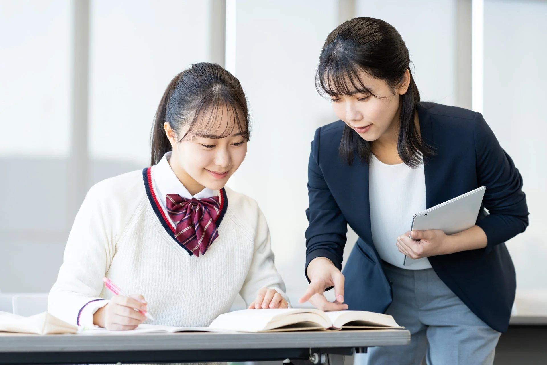 女子学生が机に向かって勉強しており、女性の先生が教科書を指差しながら指導している様子