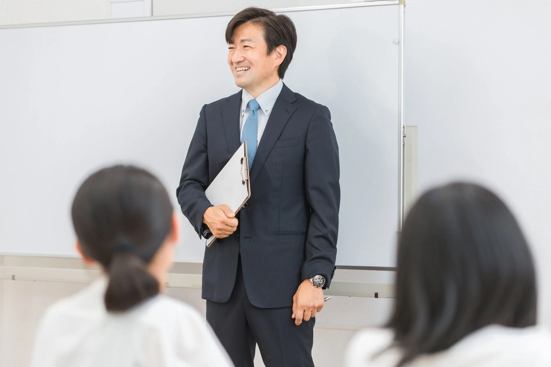 スーツ姿の男性が笑顔でホワイトボードの前に立っており、講義やプレゼンテーションを行っている場面