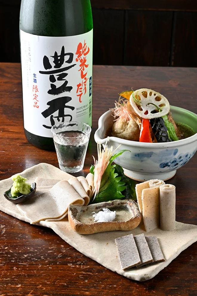 「純米しぼりたて豊盃」の日本酒と、和風の料理が並ぶ食卓の画像です。色鮮やかな煮物や、わさびと塩でいただくこんにゃくの刺身のような料理が美しく盛り付けられています。