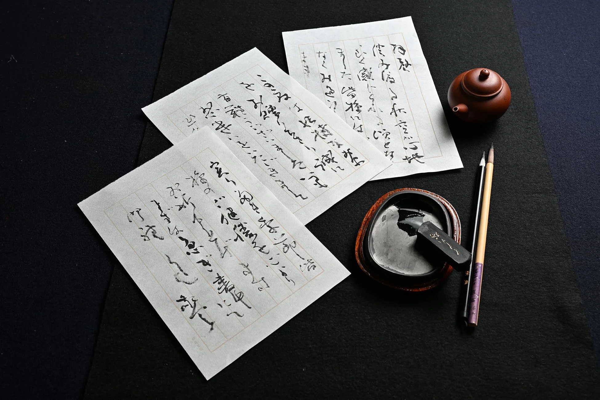 黒い背景の上に、筆と墨で書かれた古典的な日本語の書道作品が3枚置かれています。傍らには硯、墨、筆が配され、日本の伝統的な芸術と文化を感じさせる静謐な一枚です。