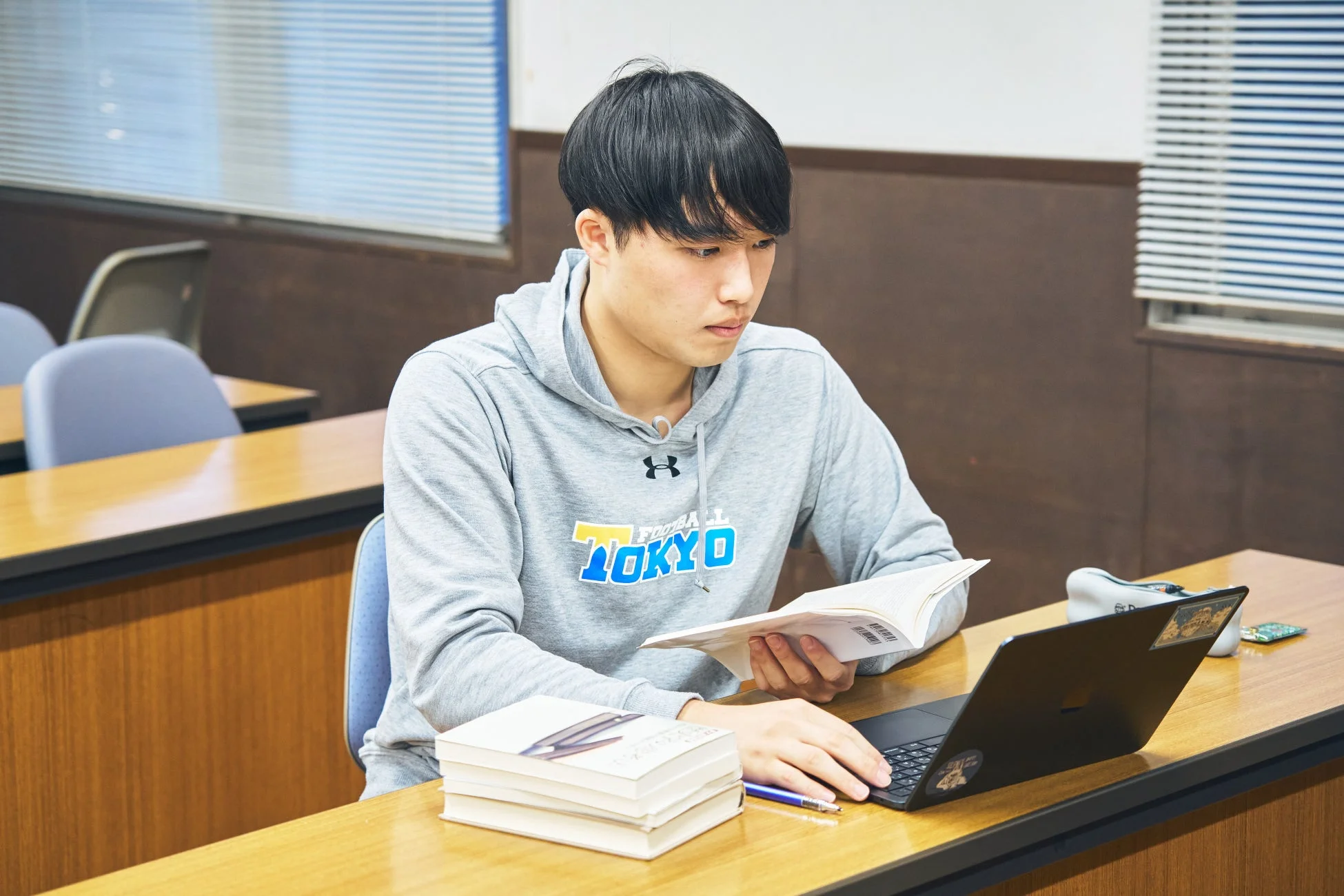 教室のような場所で、若い男性がノートパソコンと本を使って勉強している様子