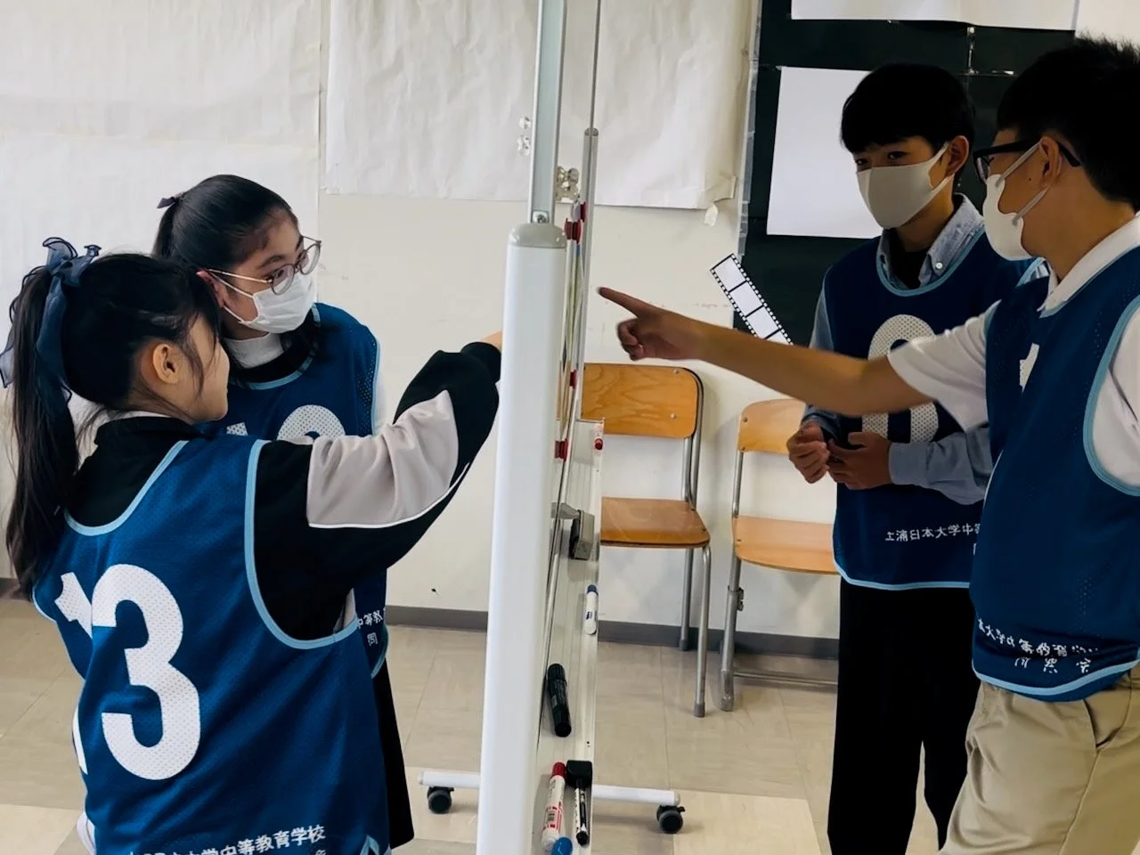教室で、マスクと青いベストを着用した4人の生徒がホワイトボードを囲んで議論しています。生徒たちはボードを指差しながら、積極的に意見を交換しているようです。