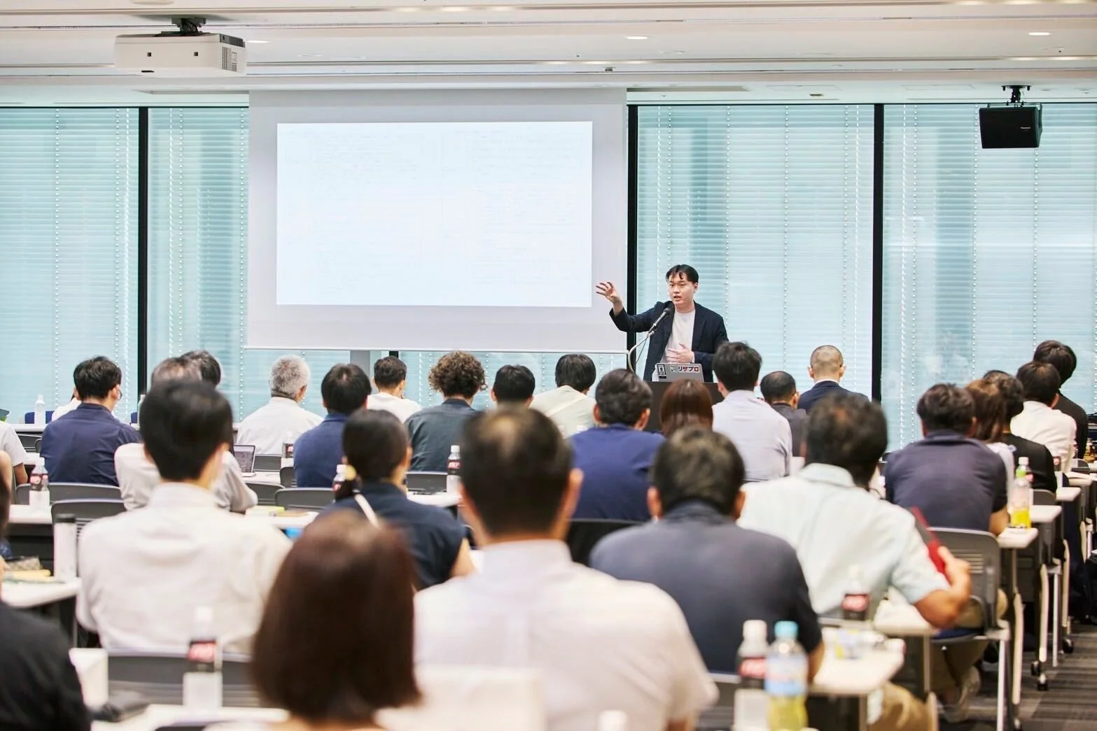 明るい会議室で、一人の男性が聴衆に向けてプレゼンテーションを行っています。壇上の男性はマイクを使い、大きなスクリーンを背景に熱心に話しており、多くの参加者が着席して耳を傾けています。