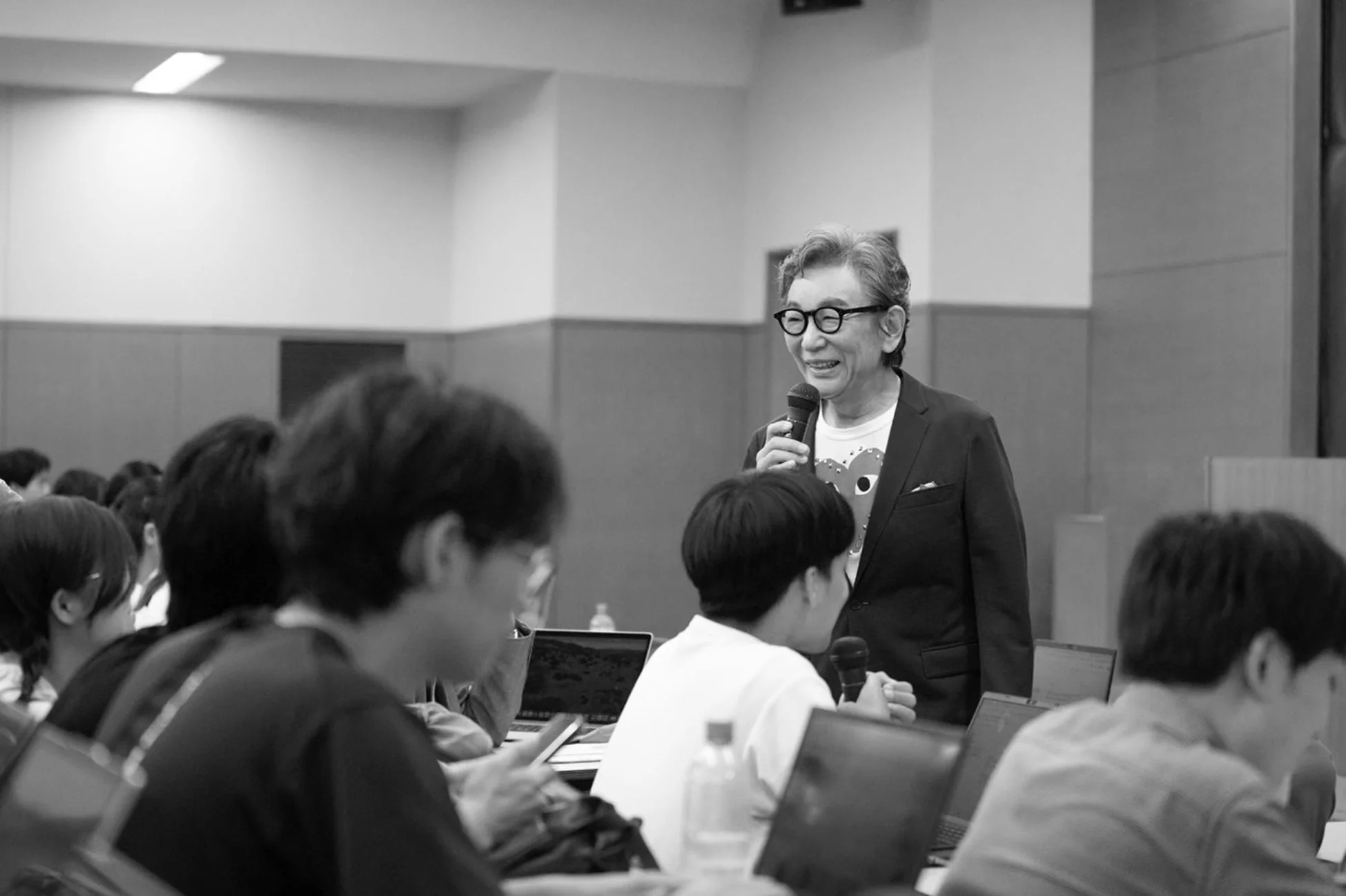 会議室のような場所で、一人の年配の男性がマイクを持って聴衆に向けて笑顔で話しているモノクロ写真です。聴衆は熱心に耳を傾け、一部の人はノートパソコンを使用しています。