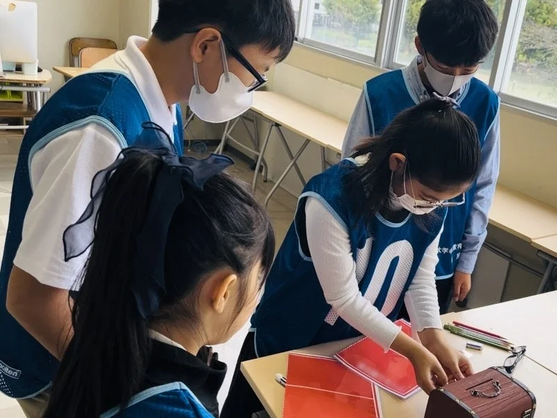マスクを着用した4人の子供たちが教室で学習活動に取り組んでいる様子。青いベストを着て、机の上で教材を使いながら協力して作業を進めています。