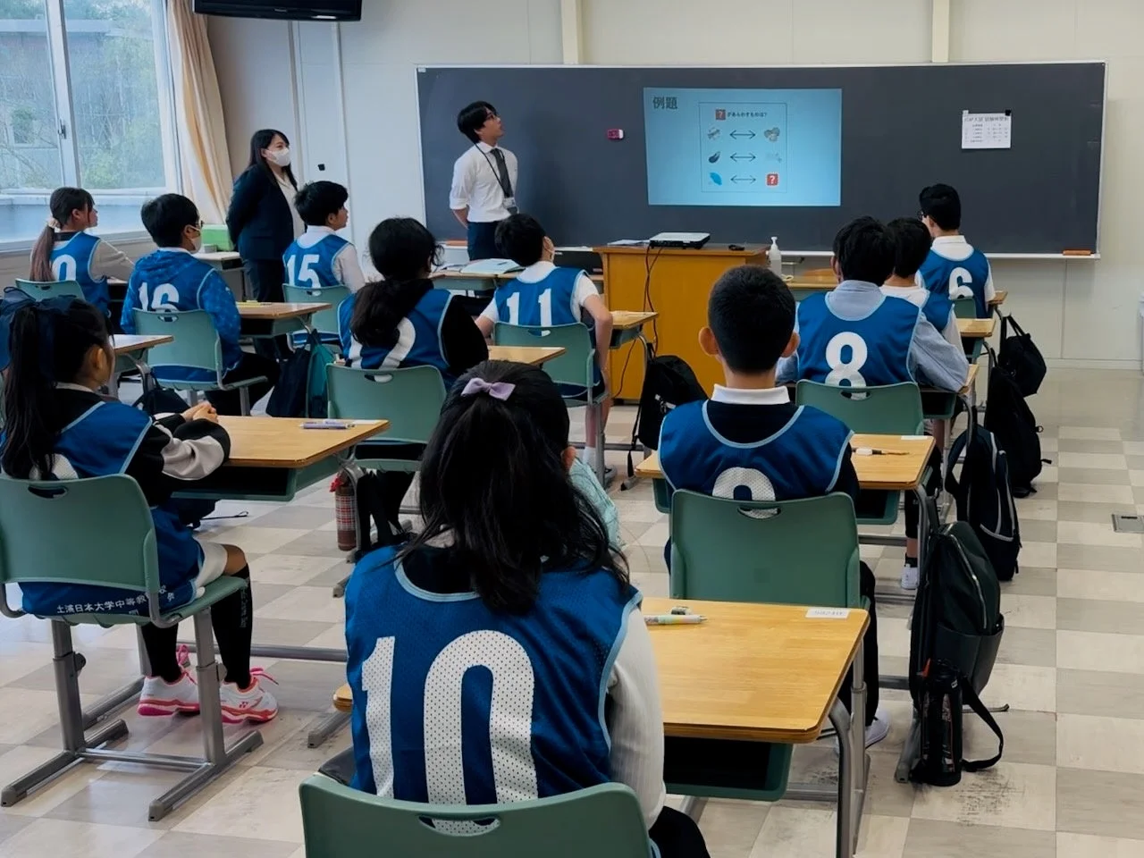 土浦日本大学中等教育学校の教室で、先生がプロジェクターを使って授業を行っています。生徒たちは番号入りの青いベストを着用し、熱心に授業を受けている様子です。