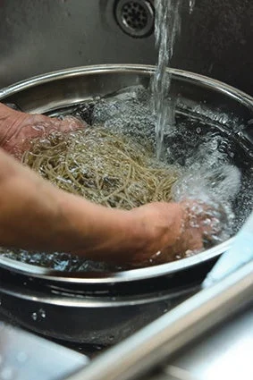 シンクの中で、人が金属製のボウルに入った麺を流水で洗っている様子。調理の下ごしらえをしている場面です。