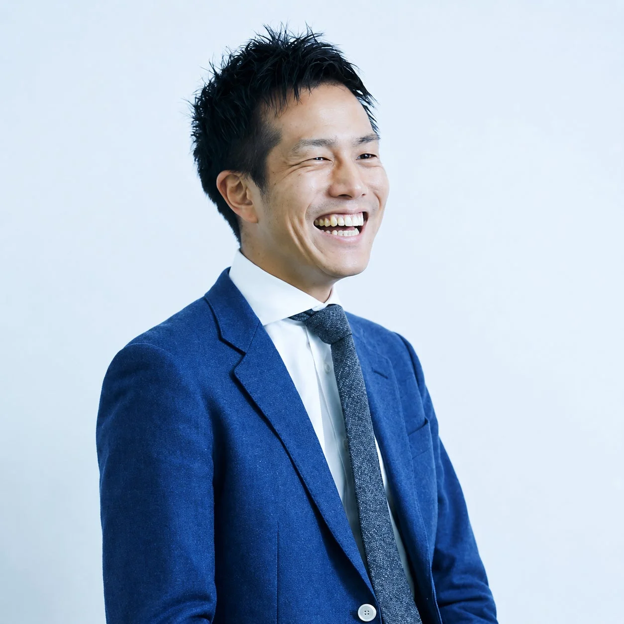 青いスーツと白いシャツを着用した日本人男性が、満面の笑みを浮かべているポートレート